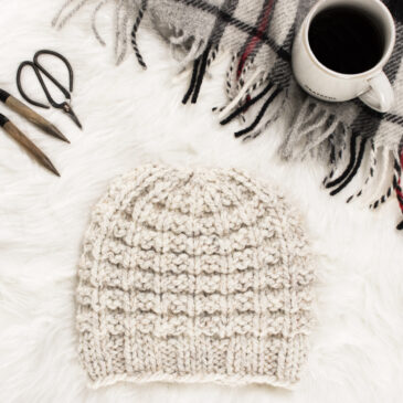 cozy scene of a hand knit chunky hat with coffee, blanket, scissors & knitting needles