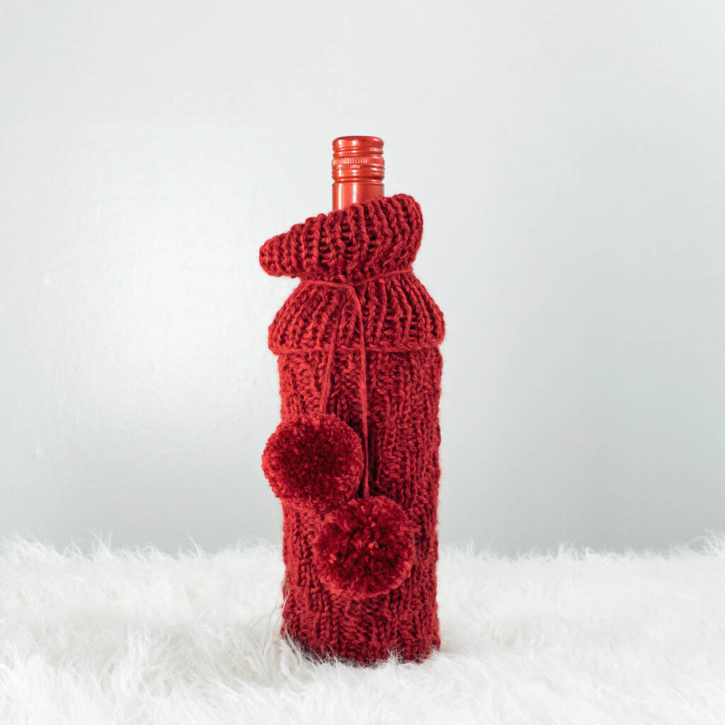 chevron knitted wine bottle cover with pom-poms displayed on a wine bottle.